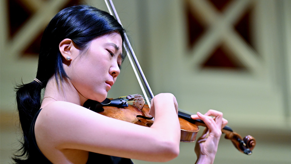 An Asian students playing the violin, surrounded by a bright environment.